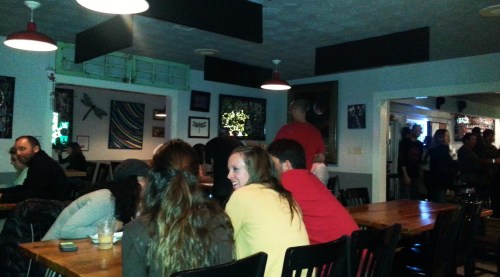 A lively bar scene with patrons seated at wooden tables, colorful artwork on the walls, and a bartender in the background.