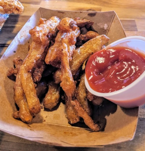 A serving of crispy fried vegetables with a side of ketchup in a brown paper tray.