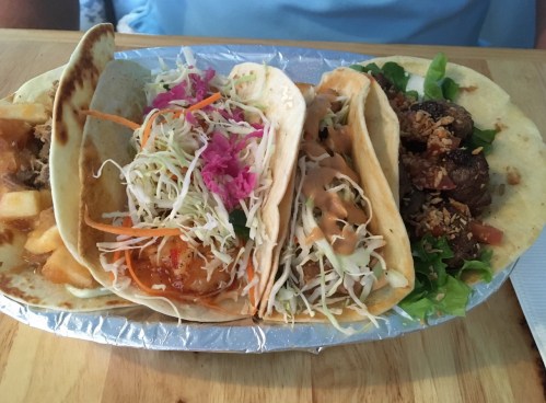 Four tacos on a tray, filled with various ingredients like cabbage, sauce, and meat, served with fresh toppings.