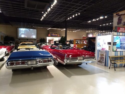 Two classic cars, a blue sedan and a red convertible, are displayed in a vintage car museum with various exhibits.