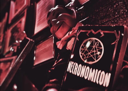 A close-up of a "Necronomicon" book cover beside a vintage puppet, with dim red lighting and framed documents in the background.
