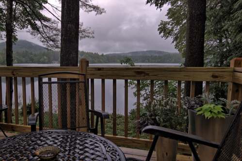 A serene lakeside view from a wooden deck, surrounded by trees and misty mountains under a cloudy sky.