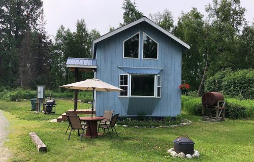 A blue two-story house surrounded by greenery, with a patio table, chairs, and a grill in the yard.