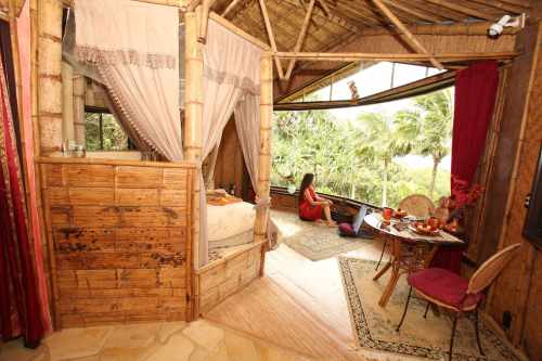 Cozy bamboo cabin interior with a woman sitting at a table, surrounded by lush greenery and natural light.
