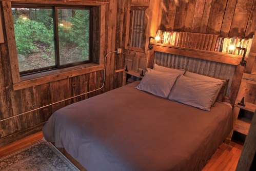 Cozy bedroom with a wooden interior, a large bed, and a window overlooking a forested area. Warm lighting adds ambiance.