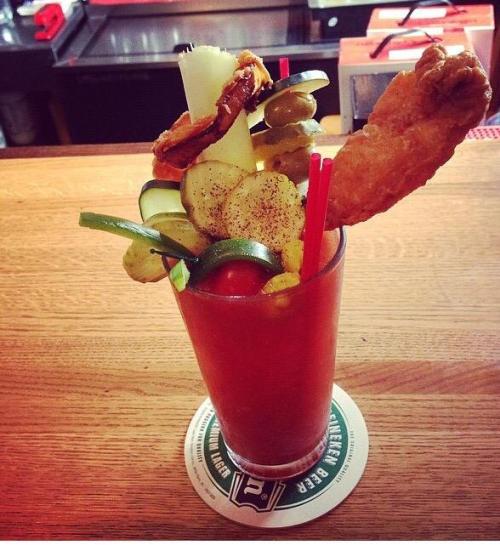 A colorful cocktail garnished with celery, pickles, olives, and a chicken wing, served in a tall glass on a wooden bar.