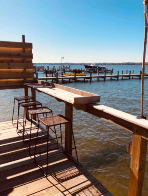A wooden deck with metal stools overlooking a calm waterway and boats docked in the distance under a clear blue sky.