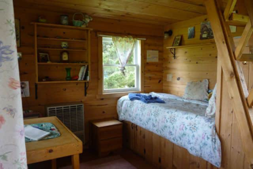 Cozy wooden cabin interior with a bed, shelves, and a small table, surrounded by nature.