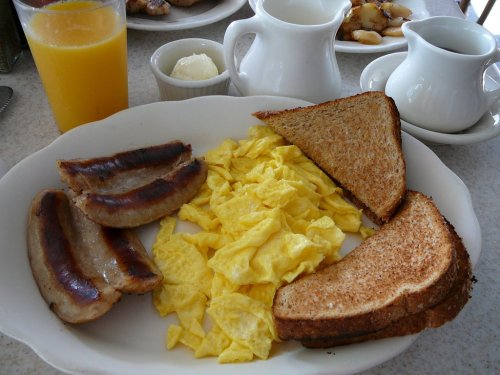 A plate with scrambled eggs, grilled sausages, and toasted bread, accompanied by orange juice and cream.