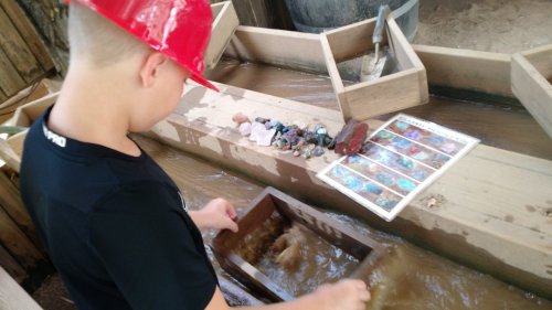 A child in a red hard hat sifts through water in a wooden trough, with colorful stones and a guide sheet nearby.