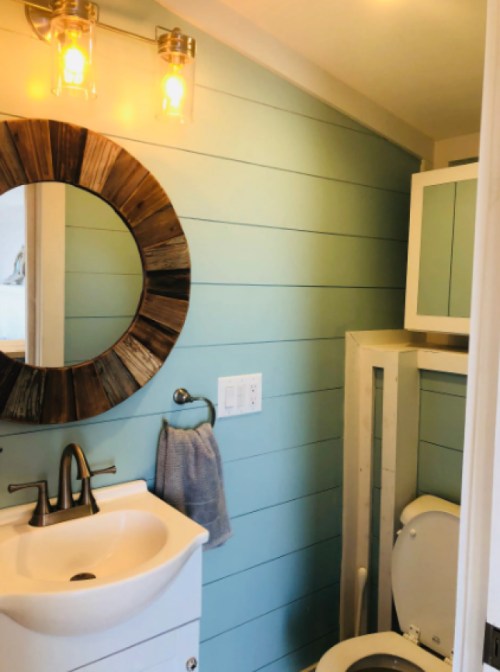 A modern bathroom with light blue shiplap walls, a round mirror, sink, and a toilet in a compact space.