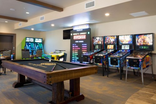A game room featuring a pool table, arcade machines, and pinball machines against a light-colored wall.