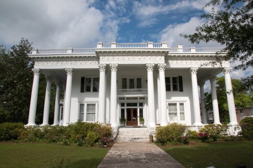 A grand white mansion with tall columns, surrounded by greenery and flowers, under a partly cloudy sky.