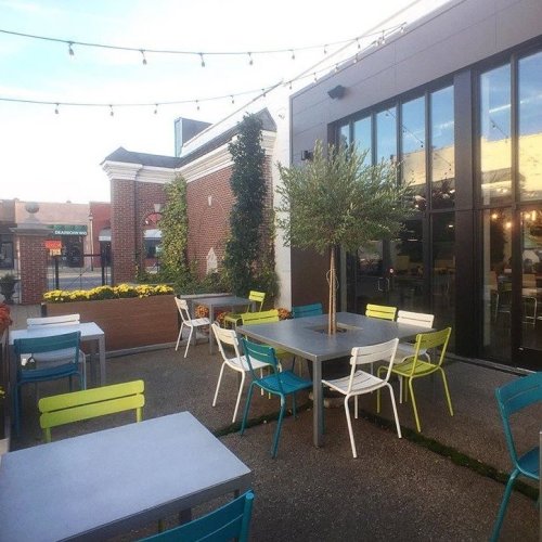 Outdoor dining area with colorful chairs, tables, and string lights, surrounded by greenery and a modern building.
