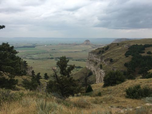 A scenic view of rolling hills and cliffs under a cloudy sky, with green vegetation and distant rock formations.