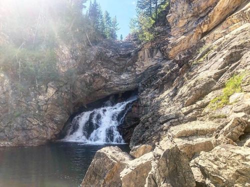 A serene waterfall cascades over rocky cliffs into a calm pool, surrounded by lush greenery and sunlight.