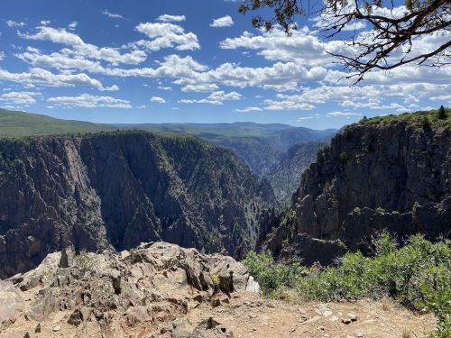 A panoramic view of a deep canyon surrounded by rugged cliffs and lush greenery under a blue sky with fluffy clouds.