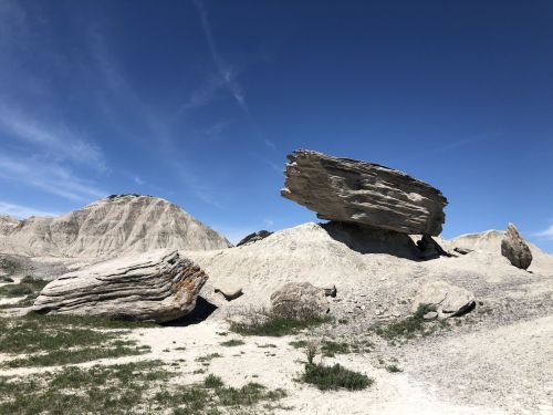 A large, flat rock balanced on a smaller rock, set against a clear blue sky and rugged terrain.