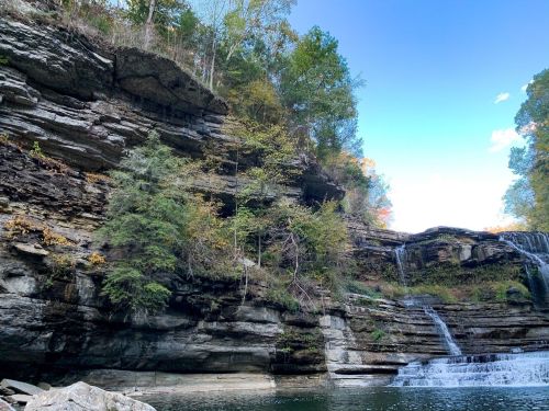 A serene waterfall cascading over rocky cliffs, surrounded by lush trees and a clear blue sky.