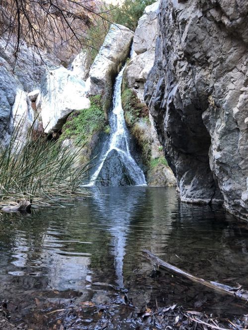 A serene waterfall cascades over rocks into a calm pool, surrounded by greenery and rugged cliffs.