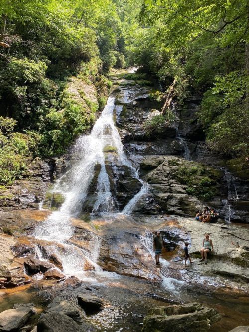 A scenic waterfall cascades down rocky terrain, surrounded by lush greenery, with people enjoying the water below.