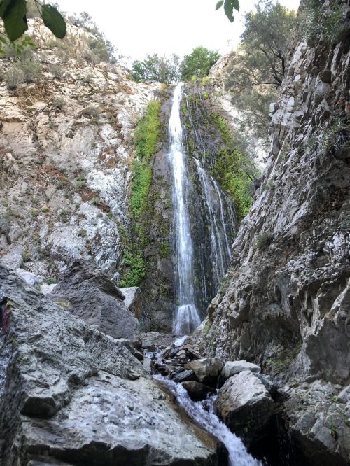 A tall waterfall cascading down rocky cliffs, surrounded by greenery and boulders in a natural landscape.