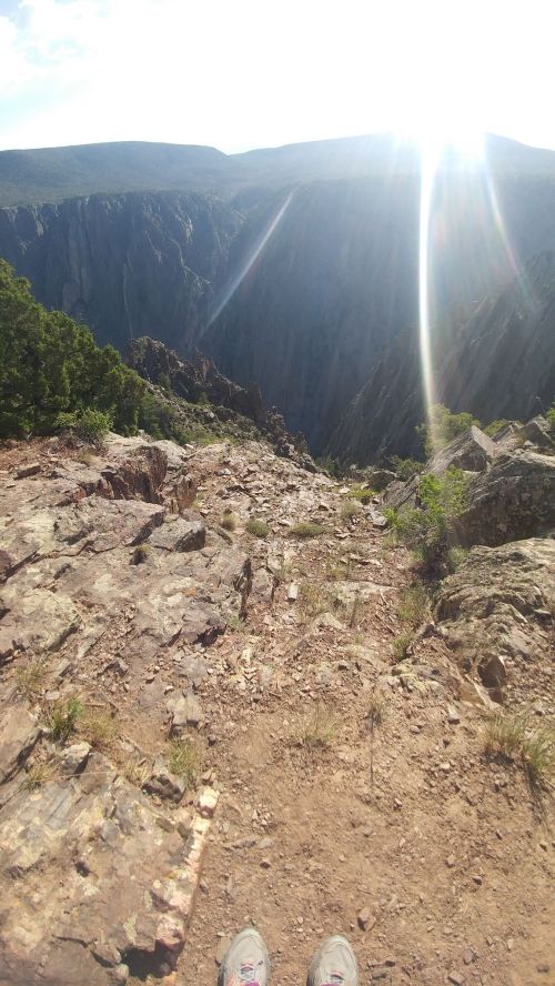 A rocky cliff edge overlooking a deep canyon, with sunlight shining brightly in the background.