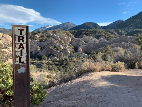 A trail sign points towards a rocky landscape with mountains in the background under a clear blue sky.