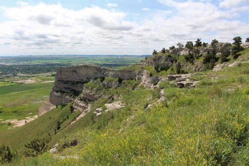 A scenic view of a rocky cliffside with green grass and wildflowers, overlooking a valley and distant hills under a cloudy sky.