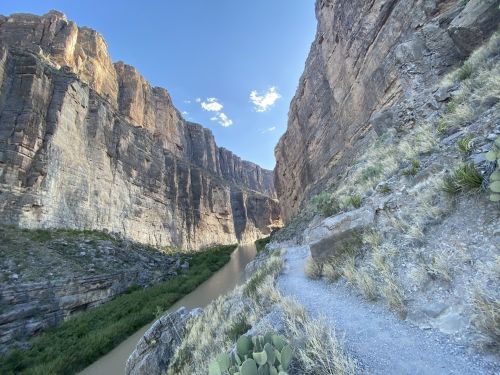A narrow canyon with steep rock walls and a river running through, surrounded by sparse vegetation and cacti.