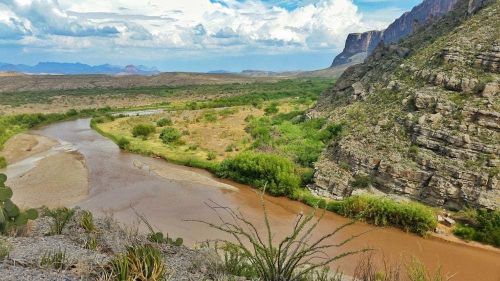 A winding river flows through a lush valley surrounded by rocky cliffs and distant mountains under a cloudy sky.