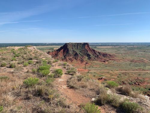 A scenic view of a red rock formation surrounded by green vegetation and open fields under a clear blue sky.