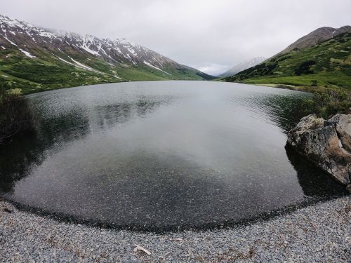 A serene lake surrounded by green hills and snow-capped mountains under a cloudy sky.