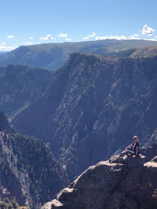 A person sits on a rocky ledge overlooking a vast mountain landscape under a clear blue sky.