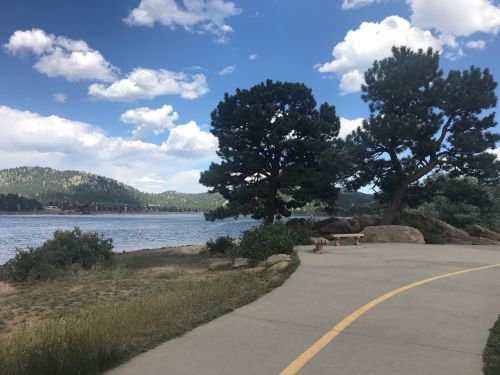A scenic lakeside path lined with trees, mountains in the background, and a partly cloudy sky.