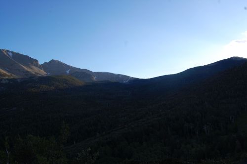 A serene mountain landscape at dusk, with peaks silhouetted against a clear blue sky and a forested valley below.