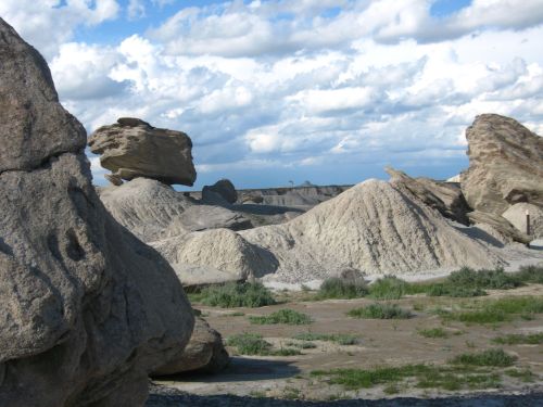 A rocky landscape with large boulders and sandy hills under a cloudy sky. Green vegetation peeks through the terrain.