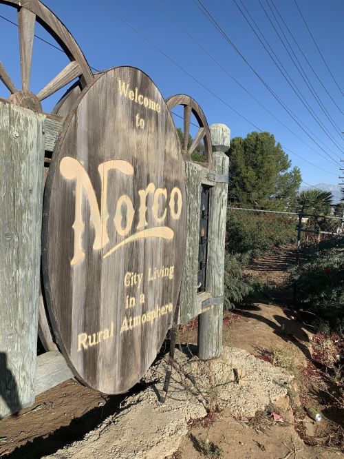 Wooden sign welcoming visitors to Norco, stating "City Living in a Rural Atmosphere," with blue sky and greenery in background.