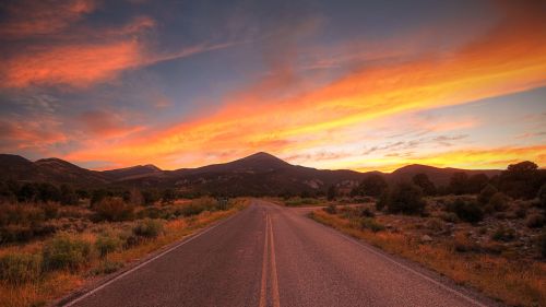 A scenic road stretches into the distance, framed by mountains and a vibrant sunset sky with orange and purple hues.
