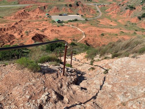 A steep, rocky descent with a railing, overlooking red hills and a parking area in the distance.