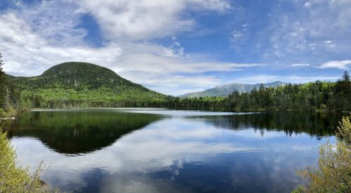 A serene lake surrounded by lush green trees and mountains, reflecting the blue sky and clouds above.