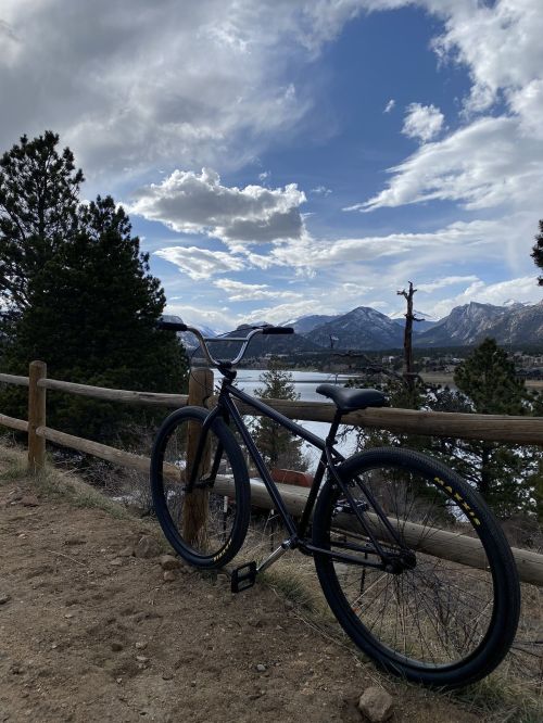 A black bicycle rests against a wooden fence, overlooking a lake and mountains under a cloudy sky.