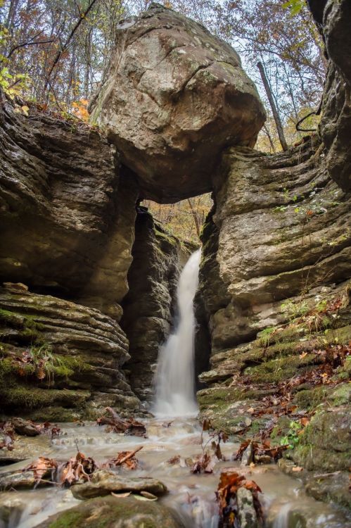 A waterfall cascades through rocky cliffs, with a large boulder perched above, surrounded by autumn foliage.