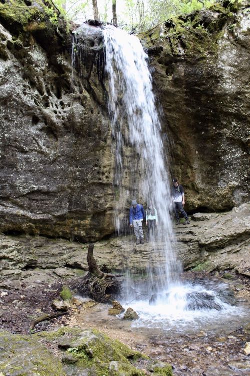 A waterfall cascades over a rocky cliff, with two people standing nearby in a lush, green forest setting.