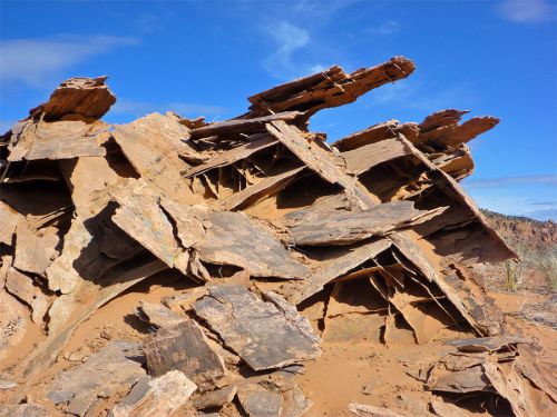 A rocky formation with layered, weathered slabs against a blue sky, surrounded by sandy terrain.