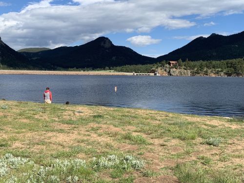 A person stands by a calm lake surrounded by mountains and greenery under a partly cloudy sky.