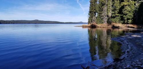 A serene lake surrounded by trees, reflecting the blue sky and distant mountains on a calm day.