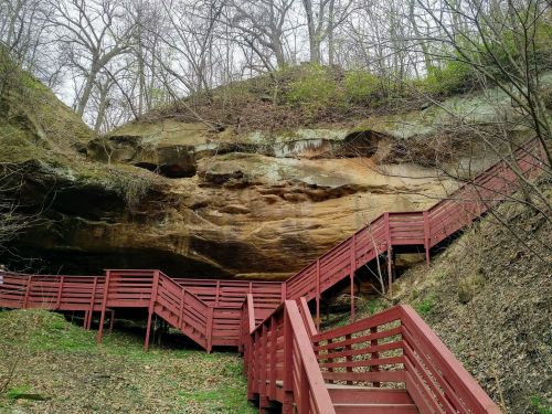 A winding red wooden staircase leads up a rocky hillside surrounded by trees and greenery.