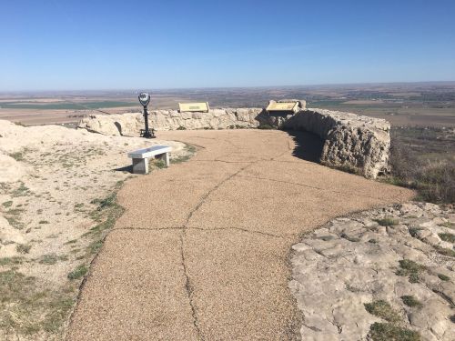 Scenic viewpoint with a path, bench, and viewing telescope overlooking a vast landscape under a clear blue sky.