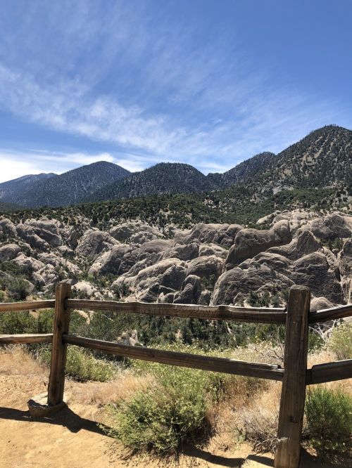 A scenic view of rocky hills and mountains under a clear blue sky, with a wooden fence in the foreground.